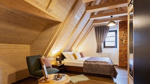 A cozy attic bedroom with a sloped ceiling, a bed with a brown bedspread, a green armchair, a small wooden table with a bowl of fruit, and a window with curtains. - B&B Plitvice Queen (Plitvicka Jezera)