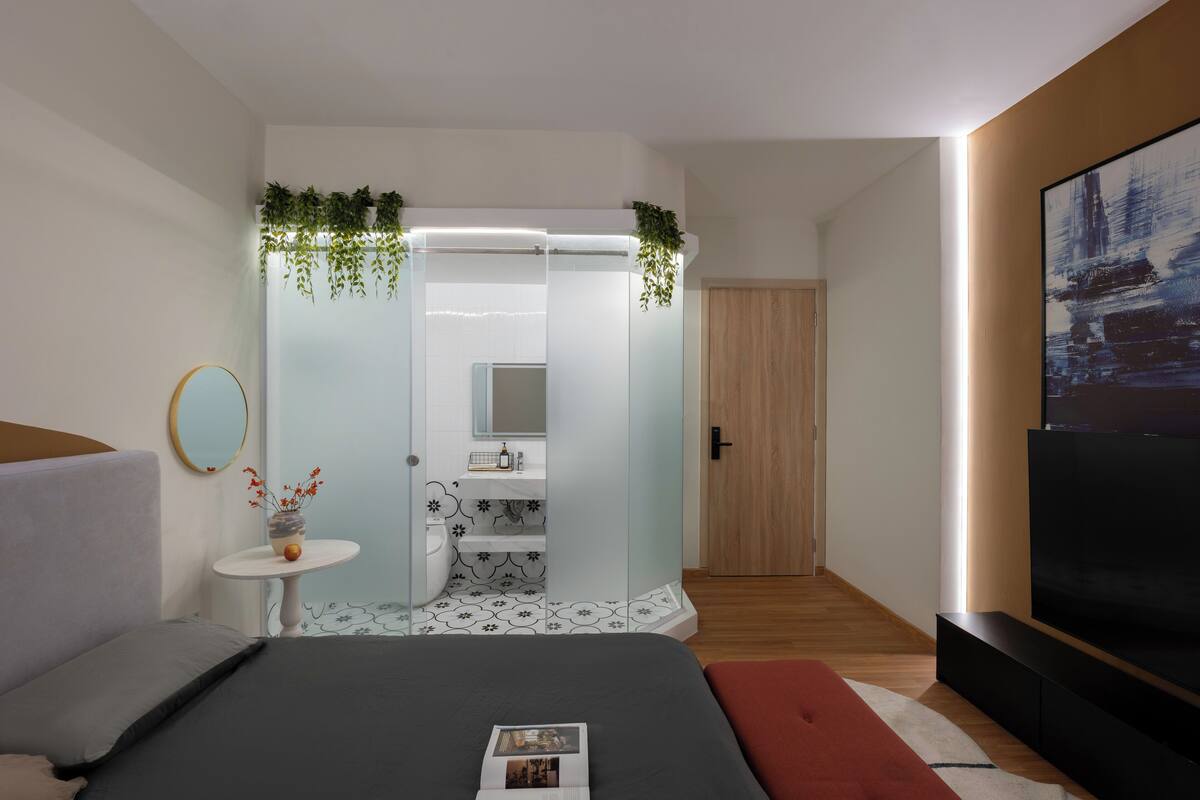 Une chambre moderne avec un lit, une petite table de chevet ornée d’un vase, un miroir, une salle de bain avec douche, et un téléviseur placé à droite.