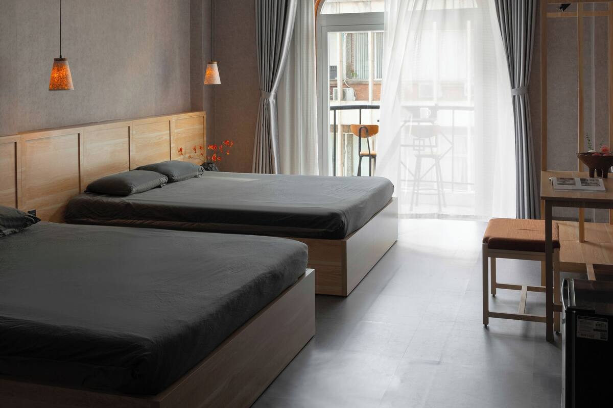 Une chambre d’hôtel moderne avec deux lits, une tête de lit en bois, une petite table et une vue sur l’extérieur grâce à une grande fenêtre agrémentée de rideaux.