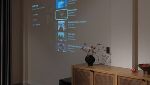 A modern living room with a wooden ceiling, a projector screen displaying a music playlist, and a wooden cabinet with a vase of flowers. - Zeus Living Central Hotel (Ho Chi Minh City)