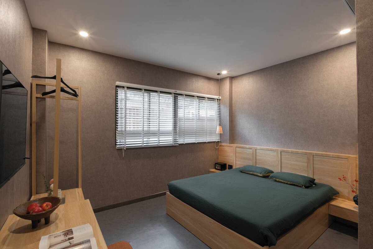 Une chambre moderne avec un lit en bois, une table de chevet, une commode, un téléviseur fixé au mur et une fenêtre avec des stores.