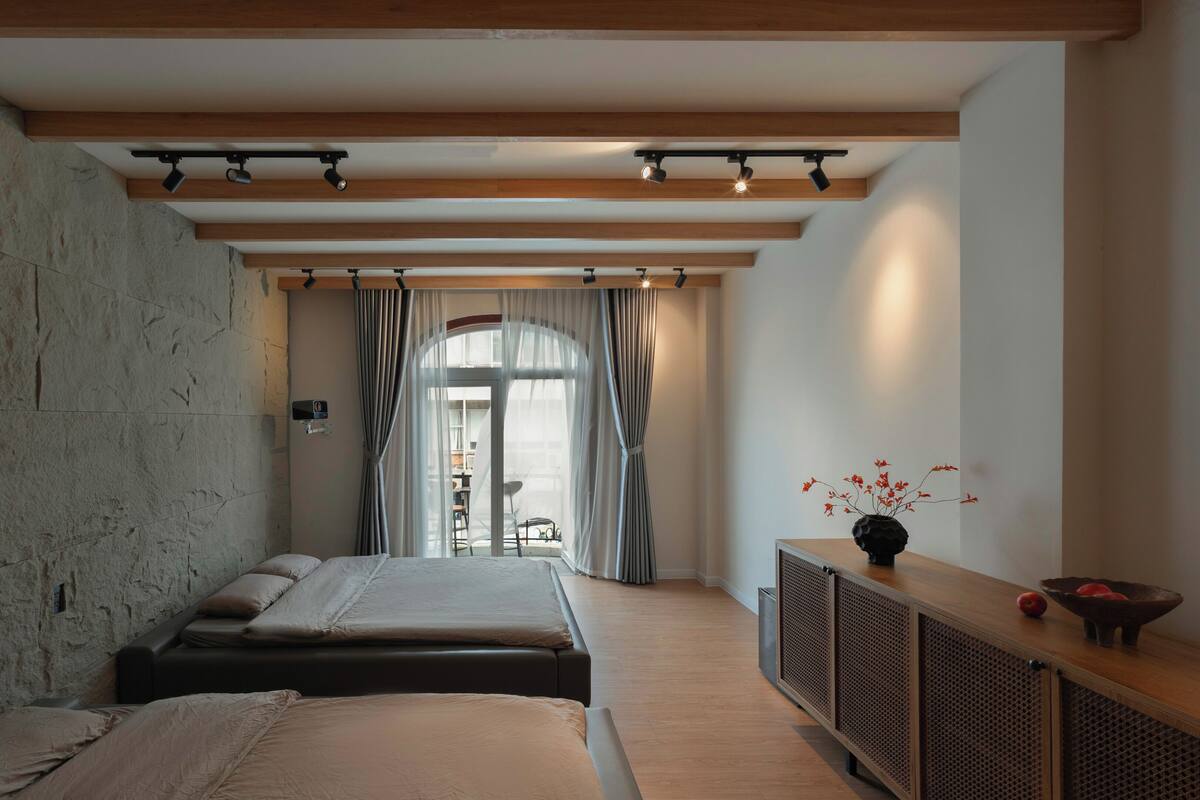 Une chambre avec un mur en pierre, un lit recouvert de plusieurs coussins, un plafond en bois et une vue sur l’extérieur à travers une fenêtre ornée de rideaux.