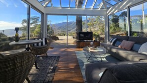 A sunlit room with a glass ceiling, a sofa, a television, and a view of mountains.