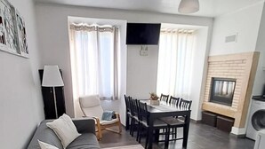 Una sala de estar moderna con un sofá, una mesa de comedor con sillas, un televisor montado en la pared y una pequeña cocina con lavadora.