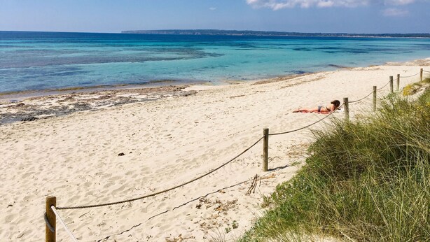 Een zandstrand met helder turquoise water, een touw afscheiding en een persoon die op het zand ligt.