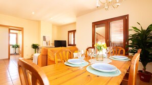 Un coin repas avec une table en bois dressée pour quatre personnes, entourée de chaises, avec un téléviseur en arrière-plan et un escalier menant à une autre pièce.