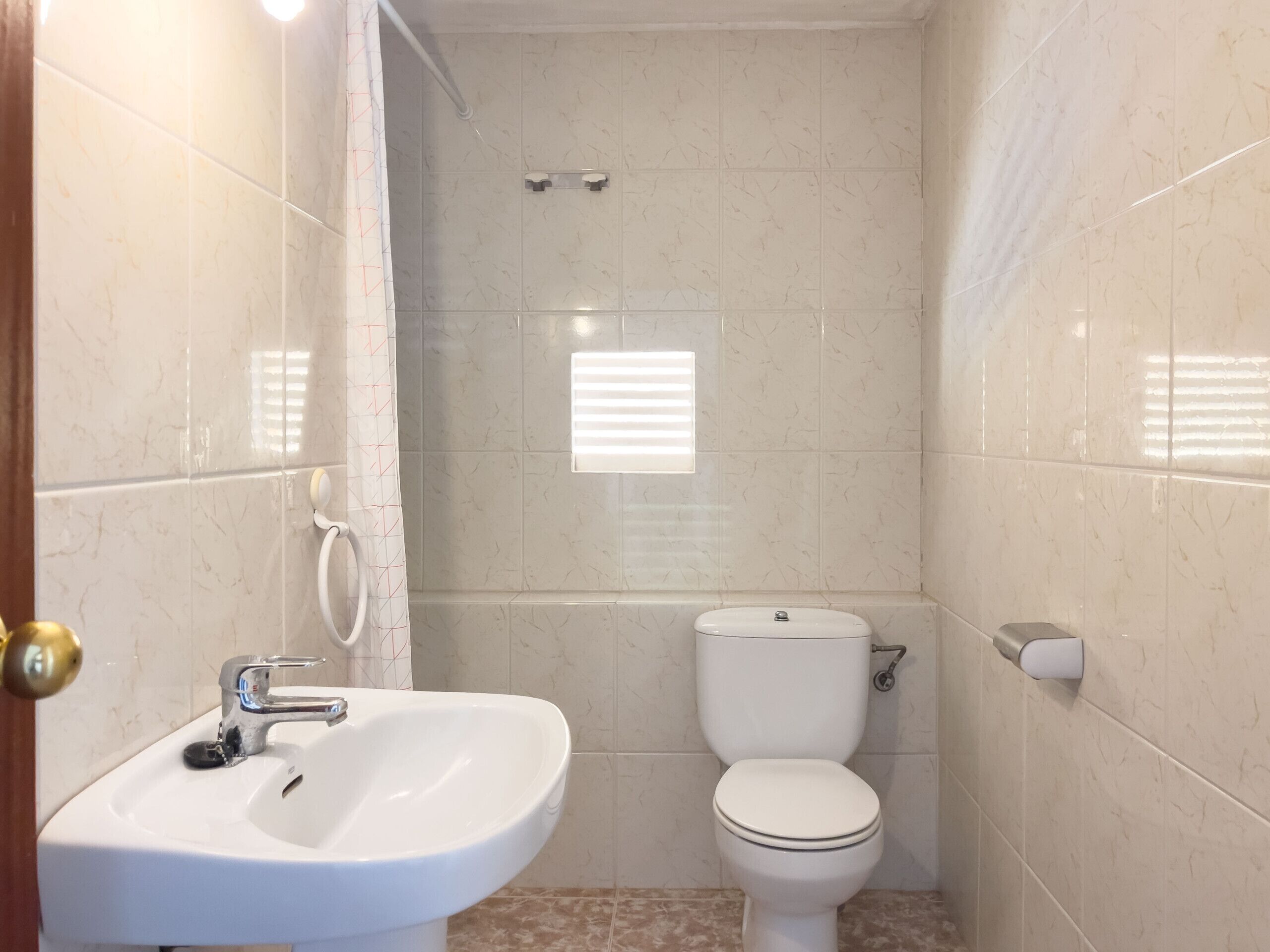 A bathroom with a sink, toilet, and shower area, featuring tiled walls and a door with a brass handle.