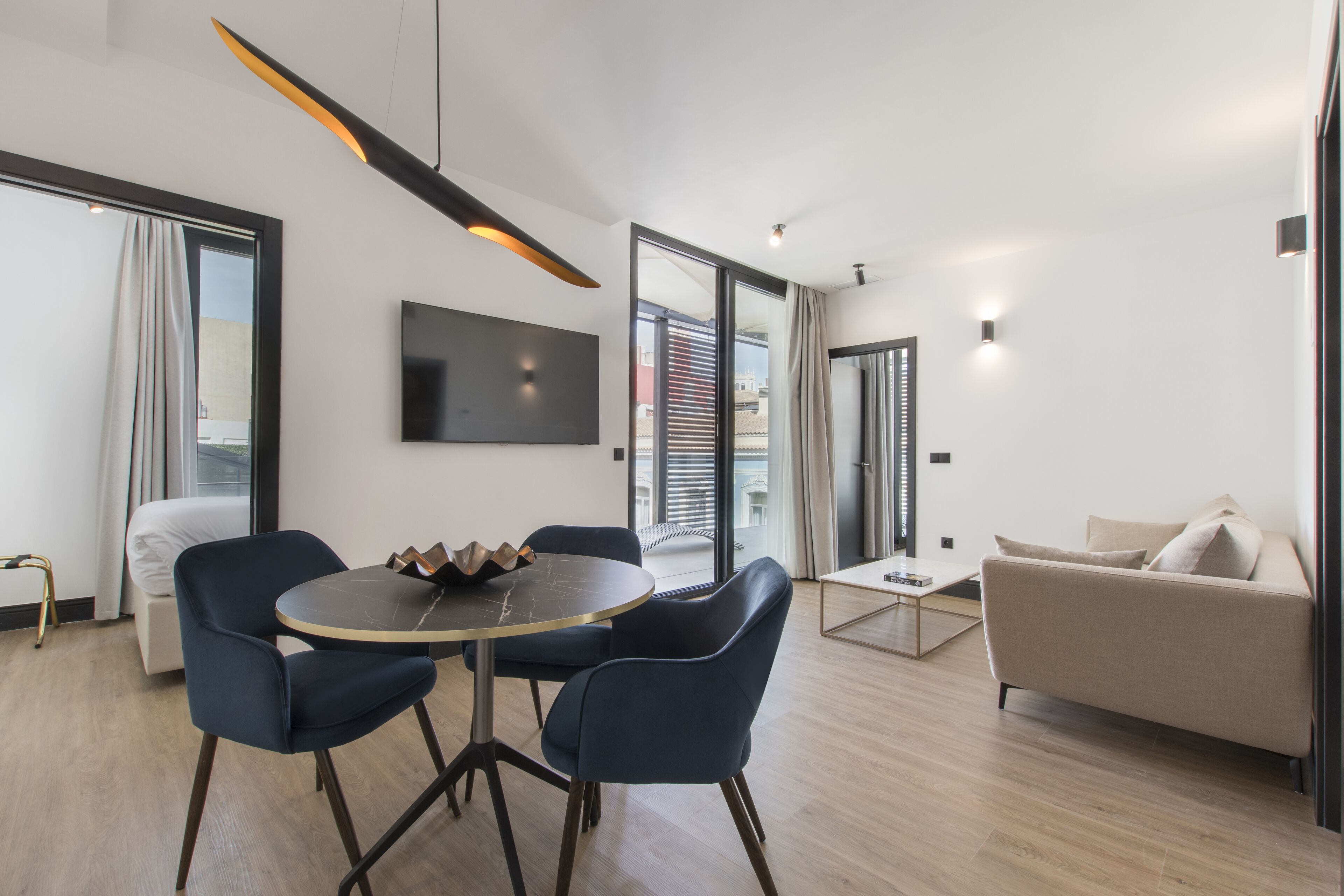 Una sala de estar moderna con una mesa redonda, dos sillas azules, un sofá beige y un televisor montado en la pared.