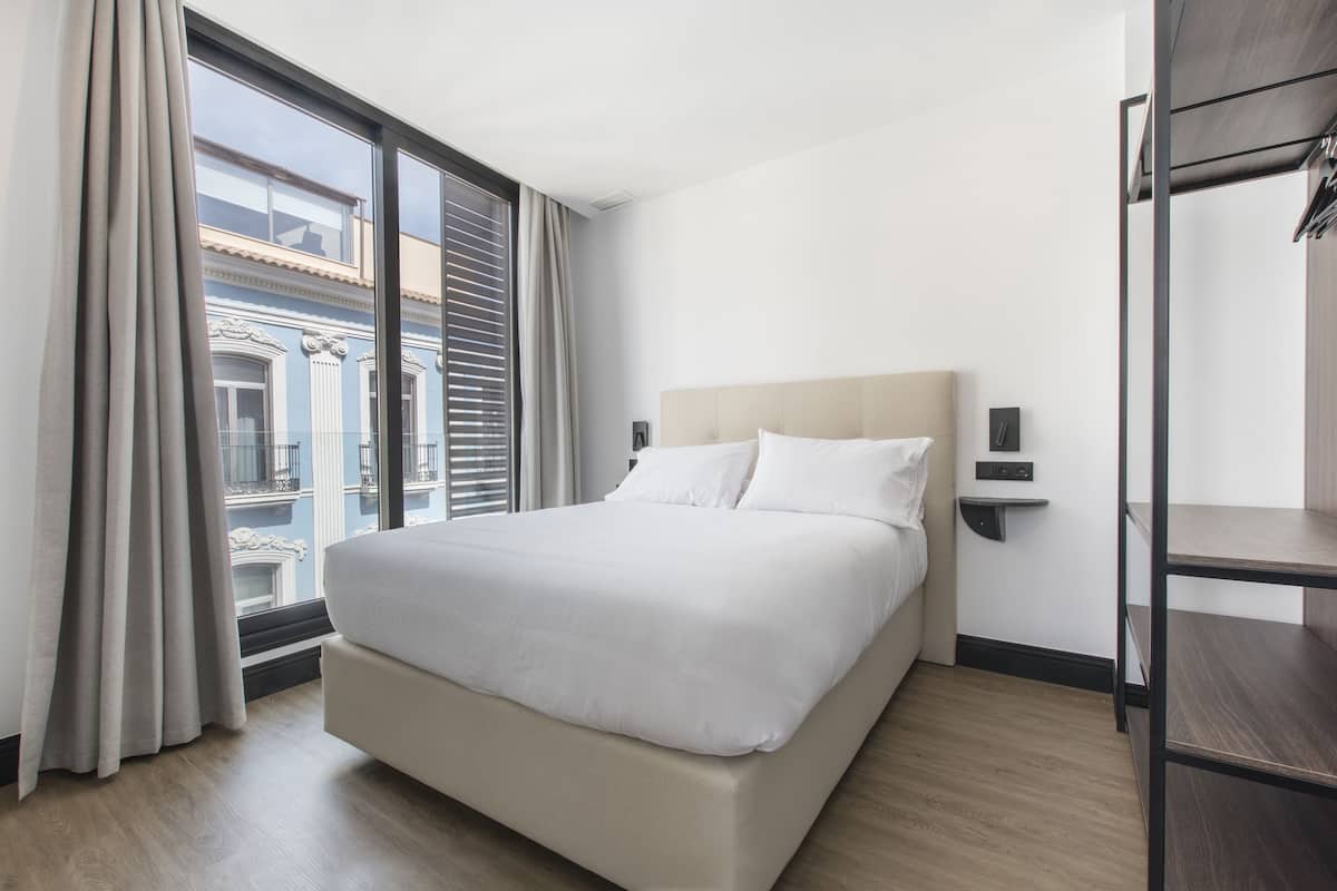 Habitación de hotel con una cama grande, una ventana con cortinas y vistas a edificios, además de piso de madera.