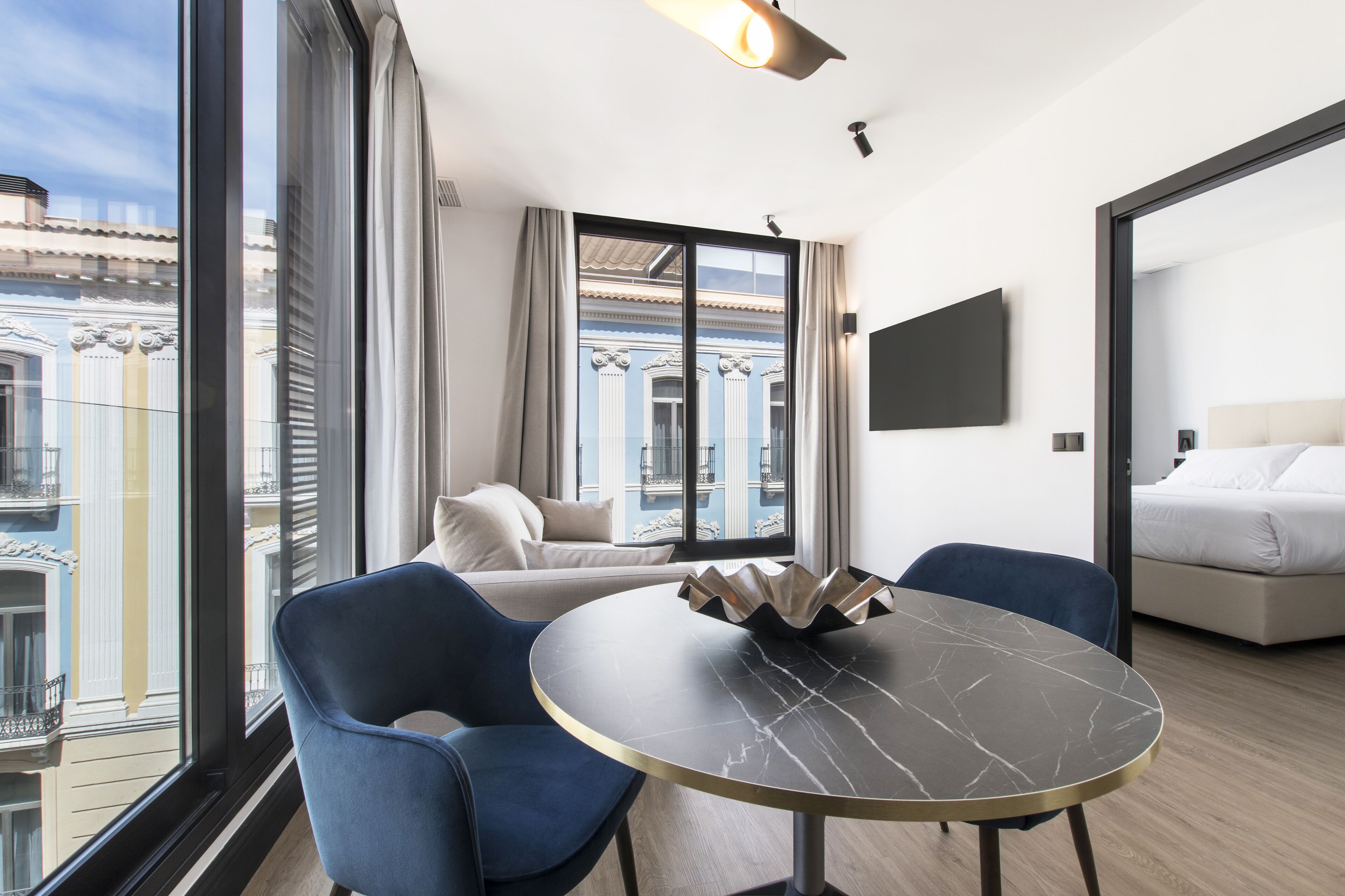 A modern hotel room with a large window, a round table with a marble top, two blue chairs, a sofa, and a bed.