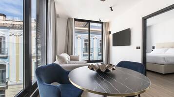 A modern hotel room with a large window, a round table with a marble top, two blue chairs, a sofa, and a bed.