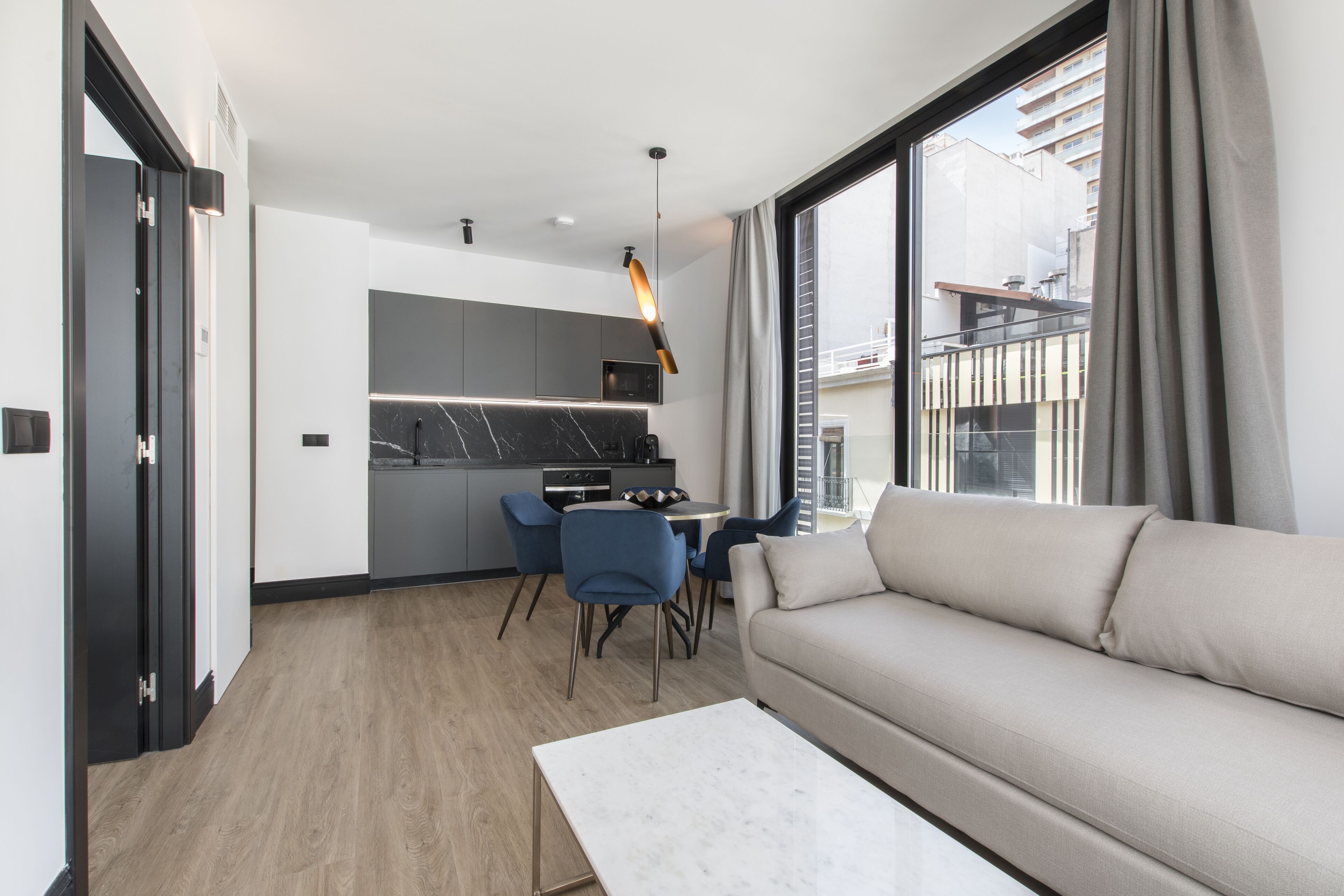 A modern living room with a sofa, a dining table with chairs, a kitchen area with a microwave, and a large window with curtains.
