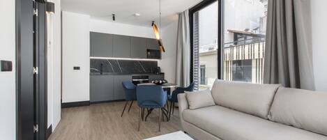 A modern living room with a sofa, a dining table with chairs, a kitchen area with a microwave, and a large window with curtains.