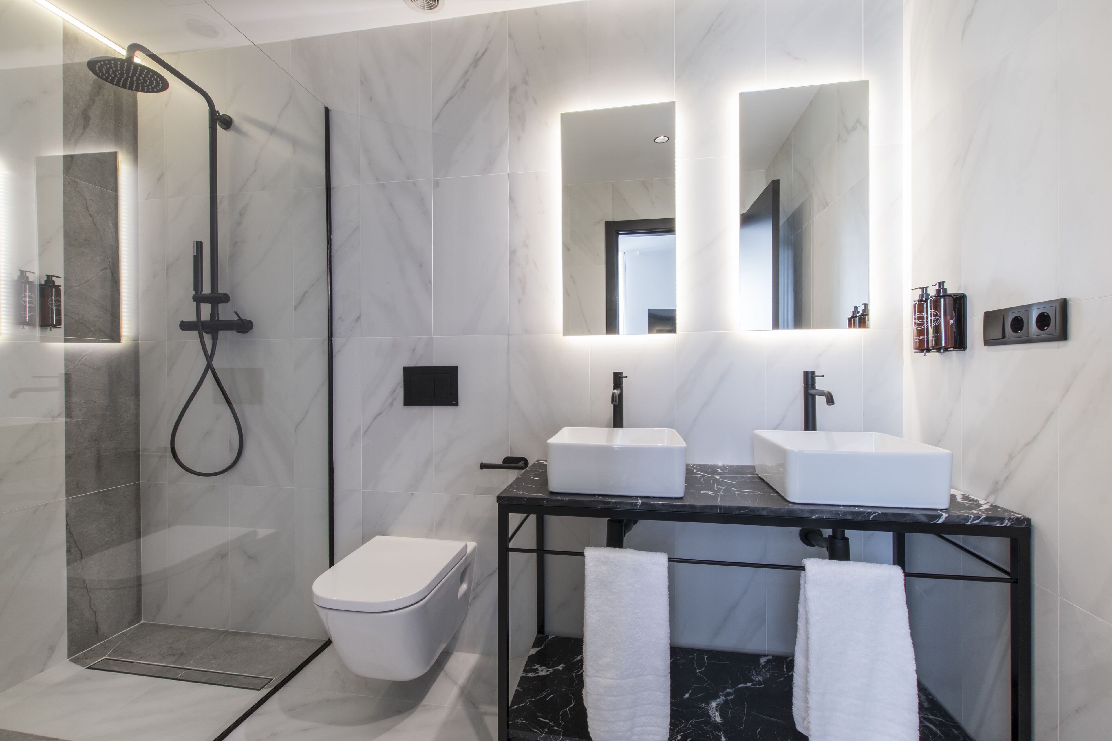 Un baño moderno con ducha envuelta en vidrio, inodoro blanco y tocador negro con dos lavabos.