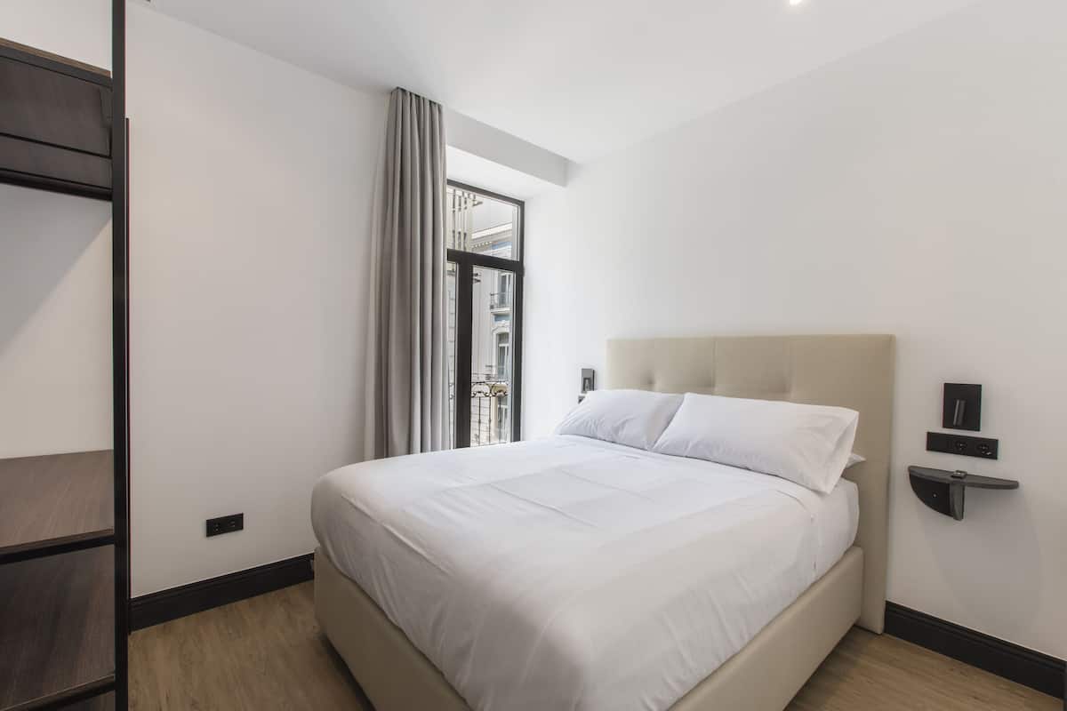 Una habitación de hotel moderna con una cama grande, una ventana con cortinas y una estantería empotrada con espejo.