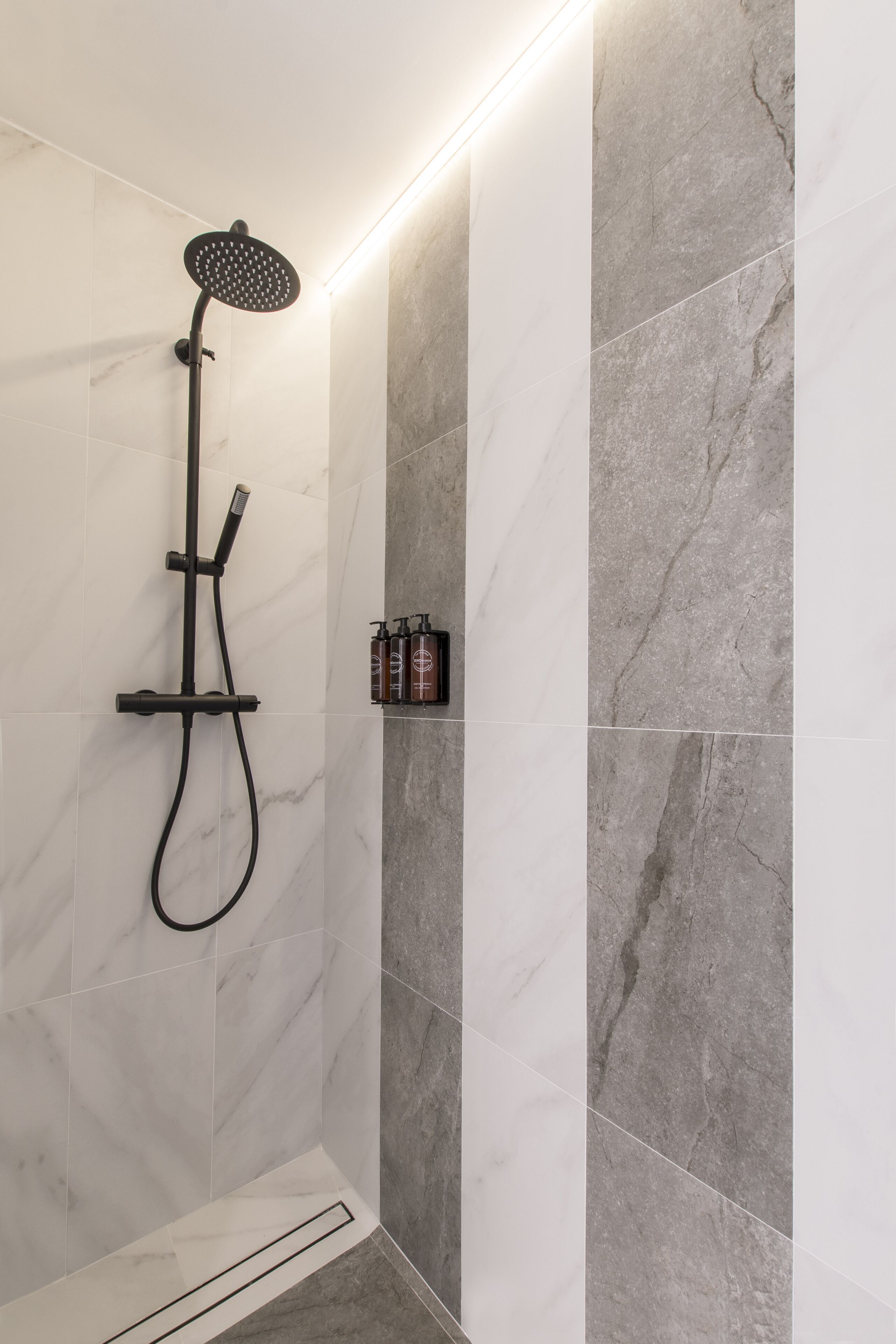 A modern bathroom with a shower, marble tiles, and a shelf with toiletries.