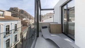 Un balcón moderno con una silla de lounge blanco y negro con estampado, una barandilla de cristal y vistas a edificios históricos y una colina al fondo.