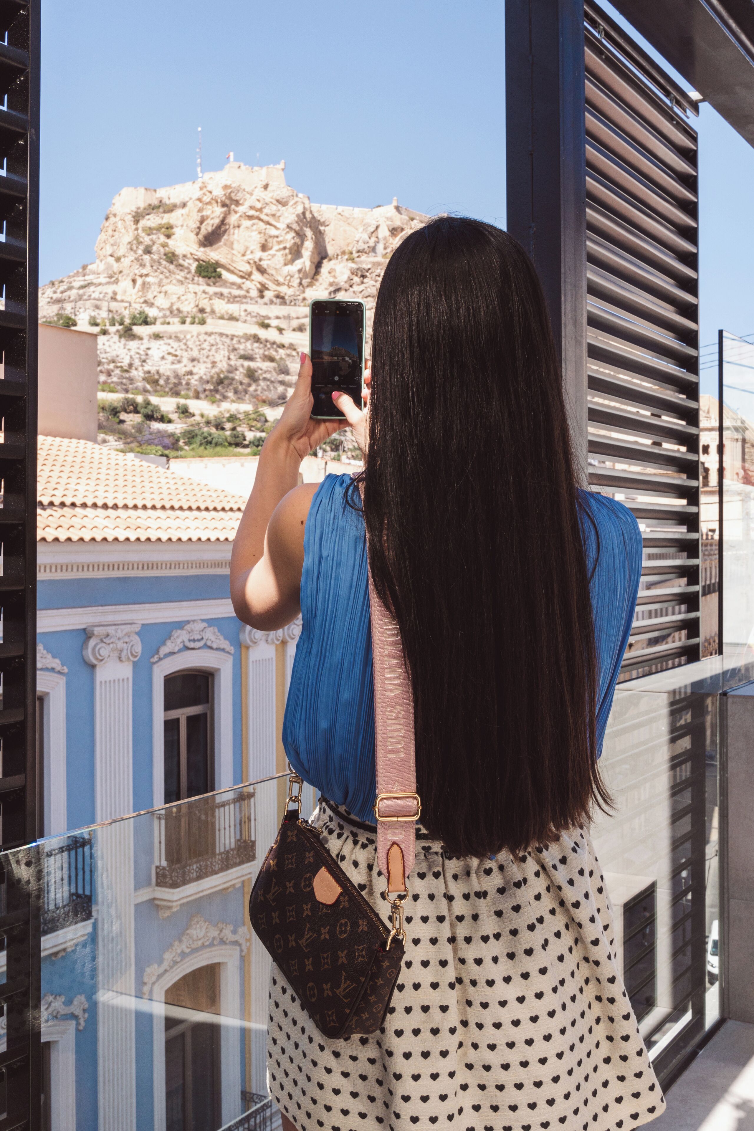 Una persona fotografiando un edificio con balcón que da a una colina rocosa.