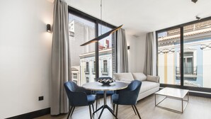 Una sala moderna con una mesa redonda, dos sillas, un sofá y una mesita de centro. Ventanas amplias ofrecen vistas a edificios con balcones.