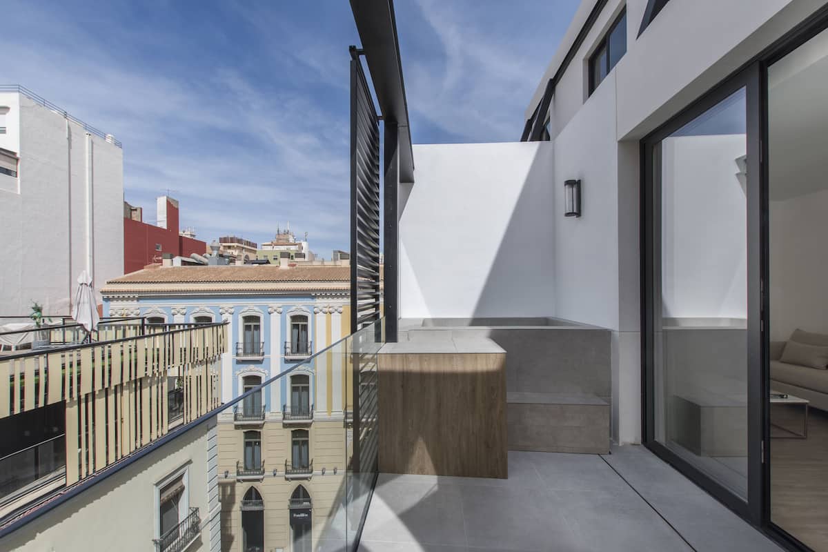 Un balcón moderno con plataforma de madera, puertas de cristal y vistas a edificios cercanos.