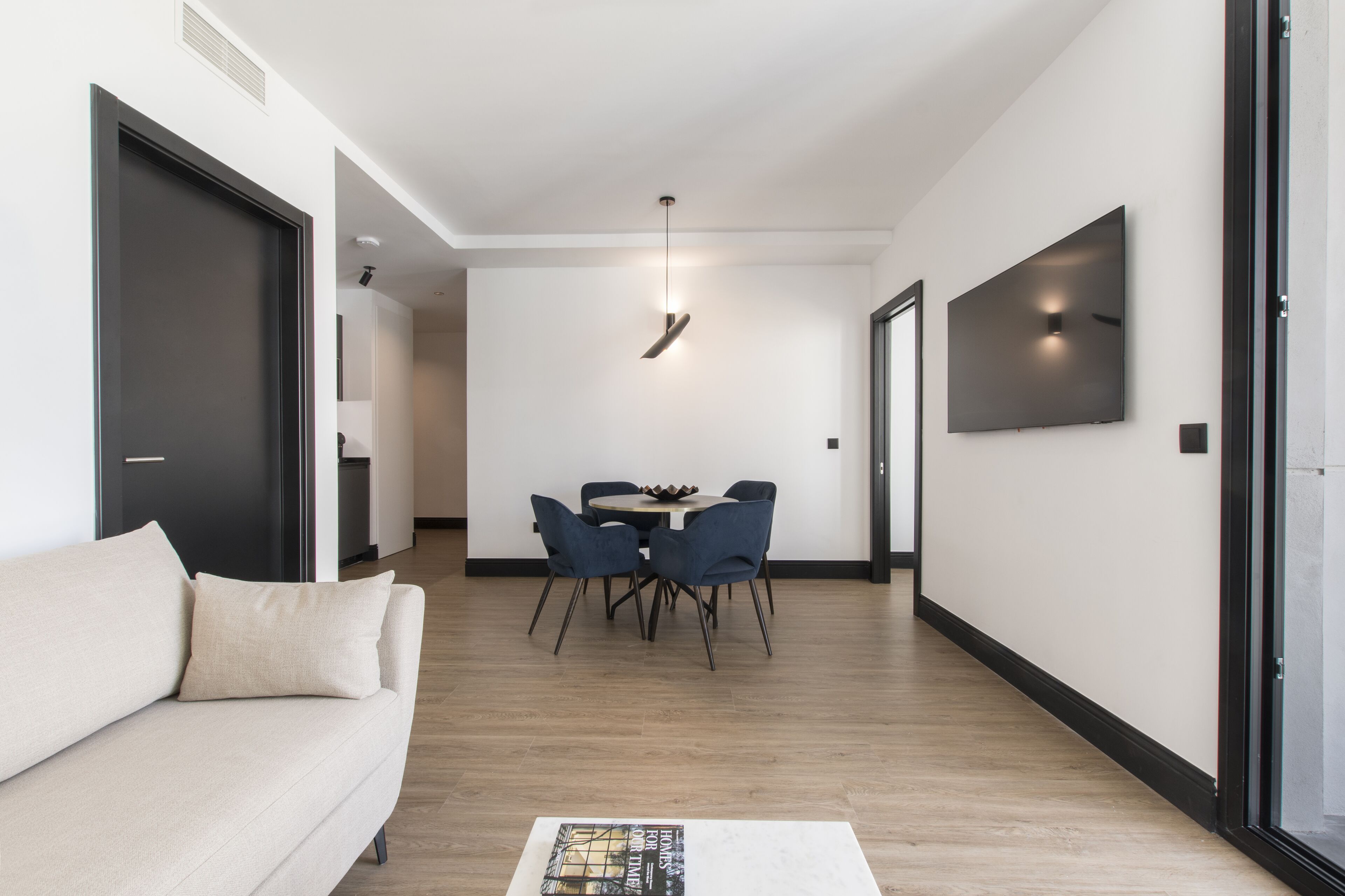 A modern living room with a sofa, a dining table with chairs, a flat-screen TV mounted on the wall, and a hallway leading to another room.
