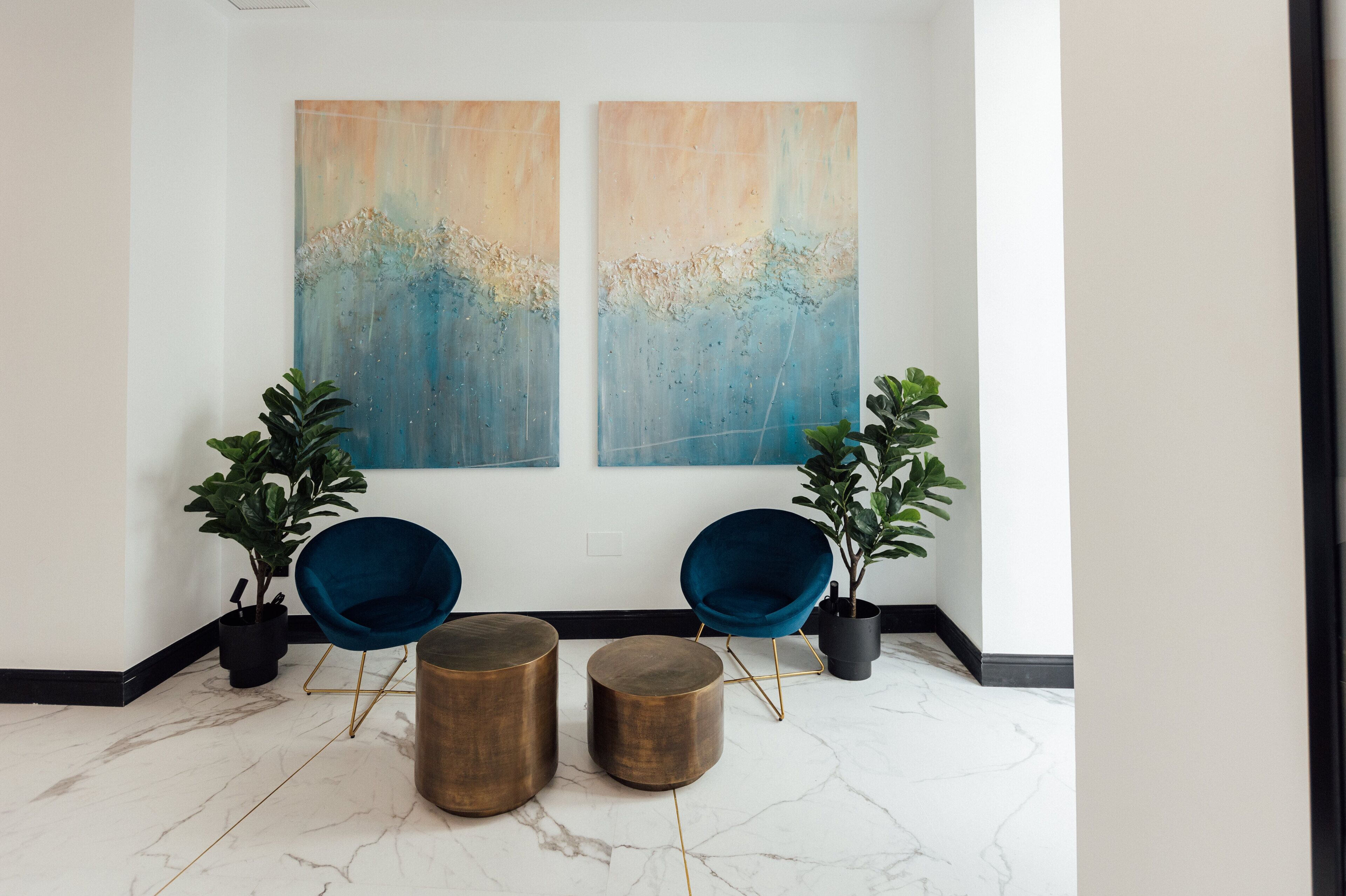 A modern lobby with two blue chairs, a marble floor, and two abstract paintings on the wall.