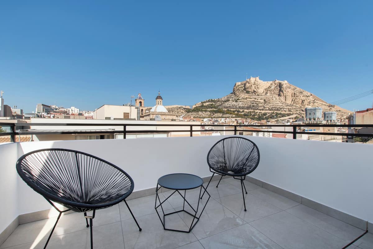 Una terraza en la azotea con dos sillas de alambre negro y una mesita pequeña, con vistas a un paisaje urbano que incluye una colina destacada y edificios históricos.