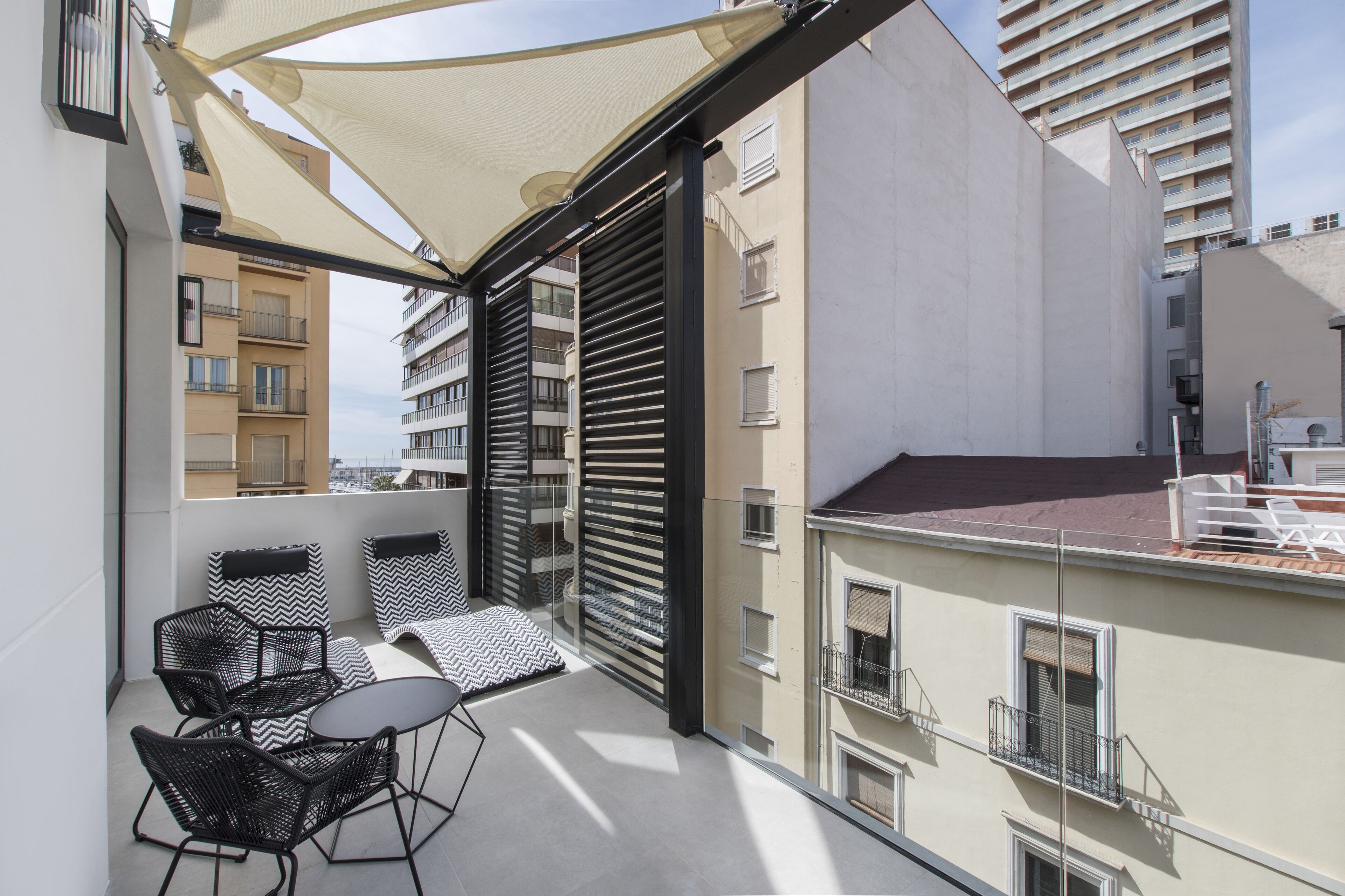 Ein Balkon mit Sitzgelegenheit, einem großen Sonnensegel und Blick auf benachbarte Gebäude.