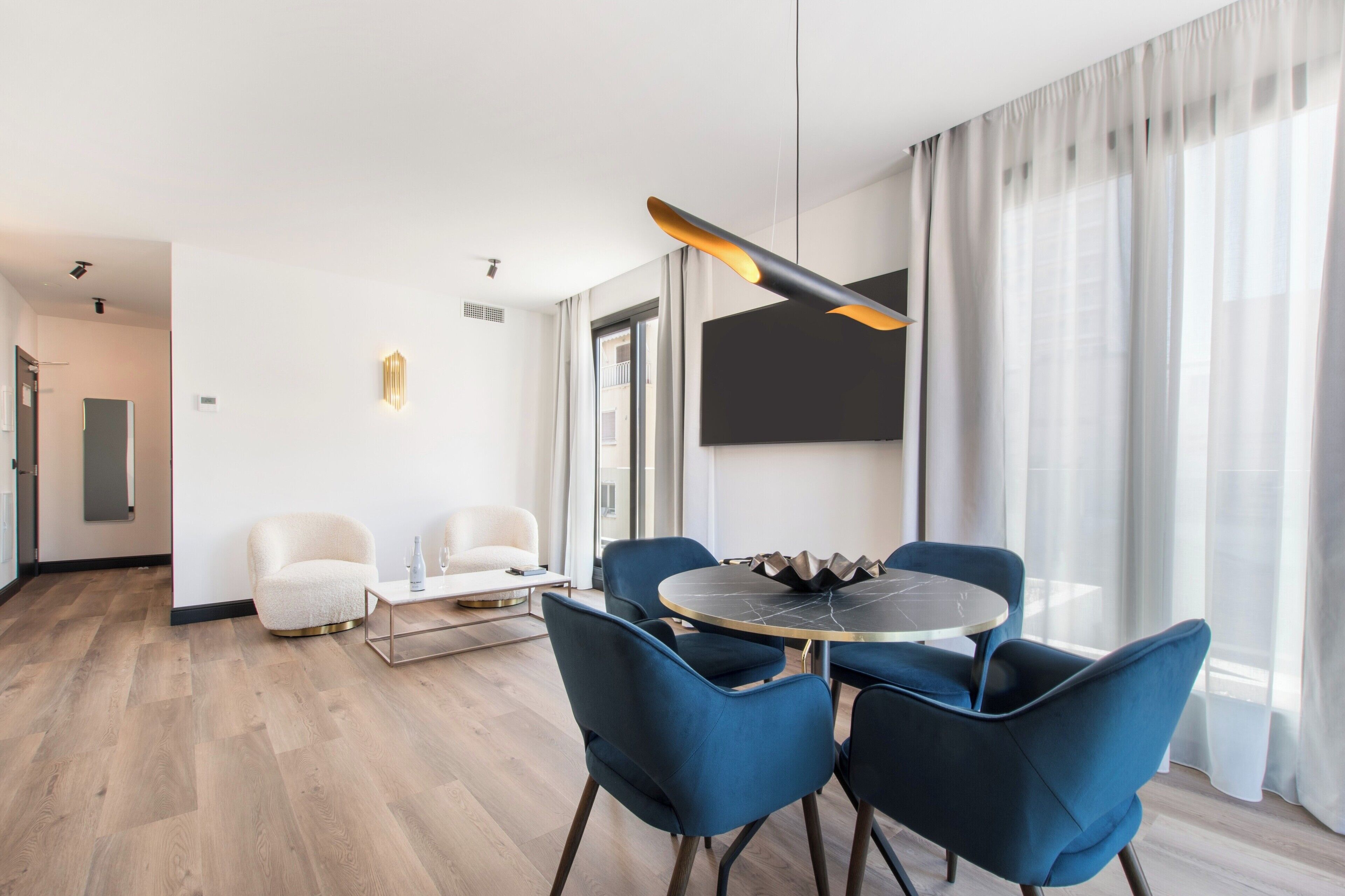 A modern hotel room with a round dining table, blue chairs, a white sofa, and a large window with sheer curtains.
