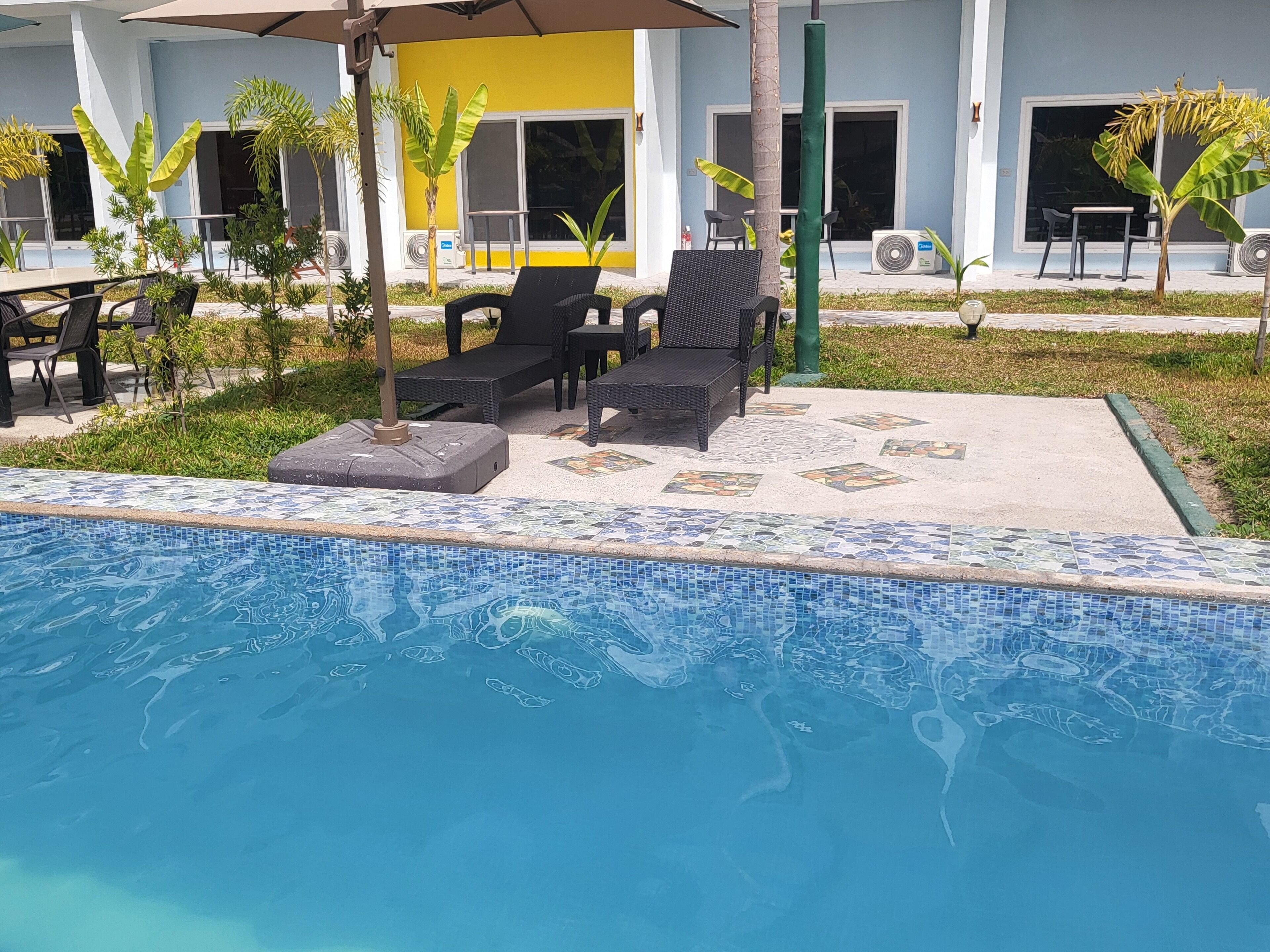 A swimming pool with lounge chairs and umbrellas, surrounded by greenery and a building with a yellow and white facade.