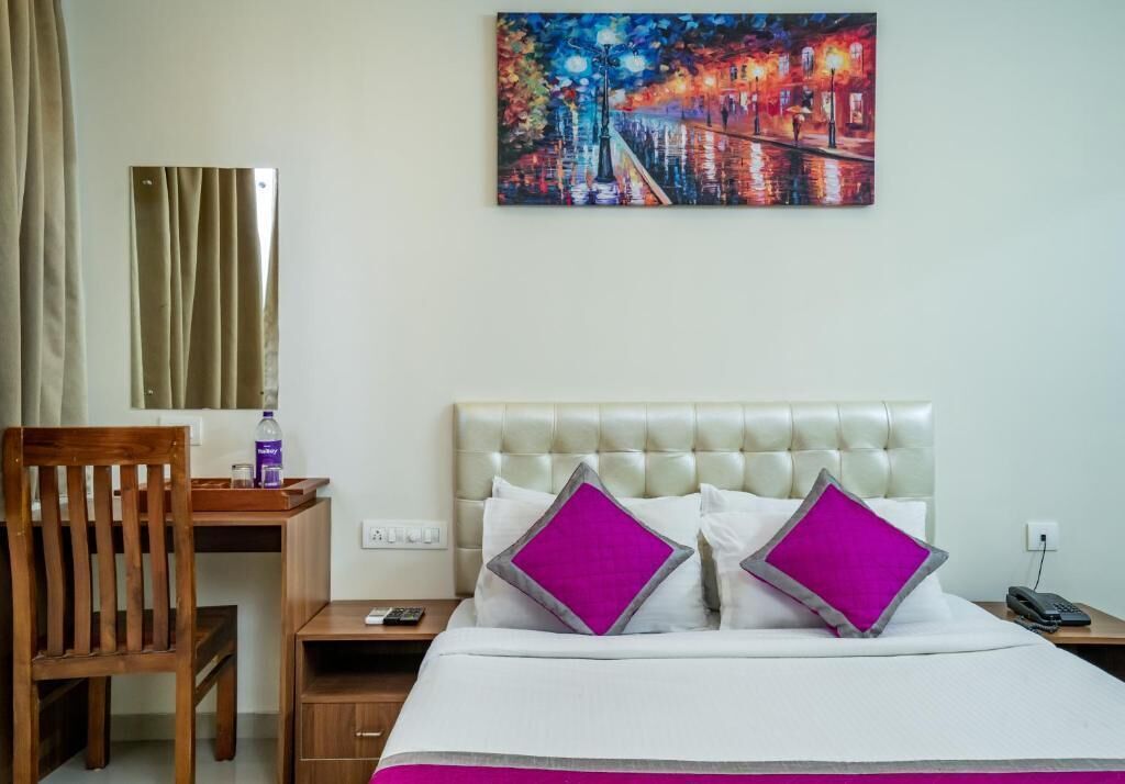 A hotel room with a bed, a wooden chair, a desk, and a painting on the wall.