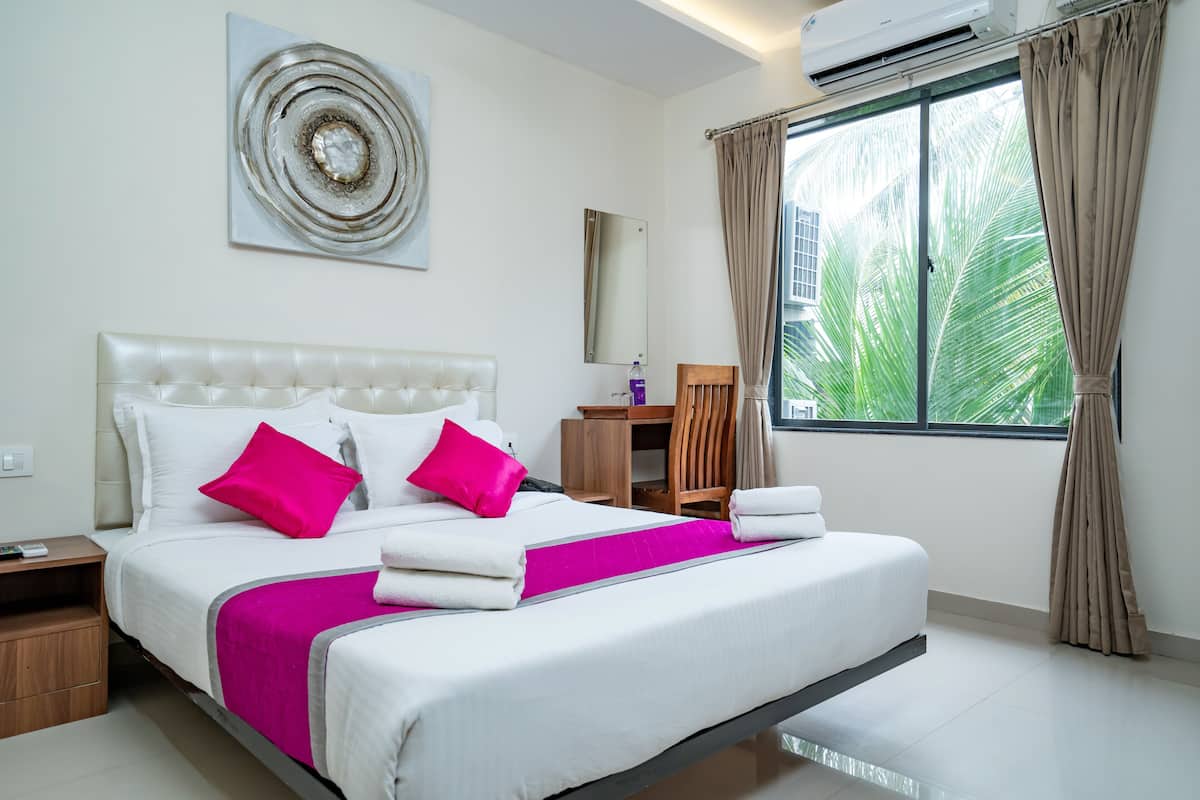 A neatly made bed with white linens and pink pillows, a bedside table with a remote control, a wooden nightstand, a mirror on the wall, and a window with curtains.