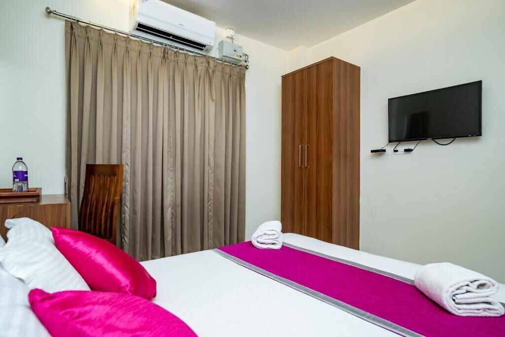 A hotel room with a bed, a TV mounted on the wall, and an air conditioning unit above the window.