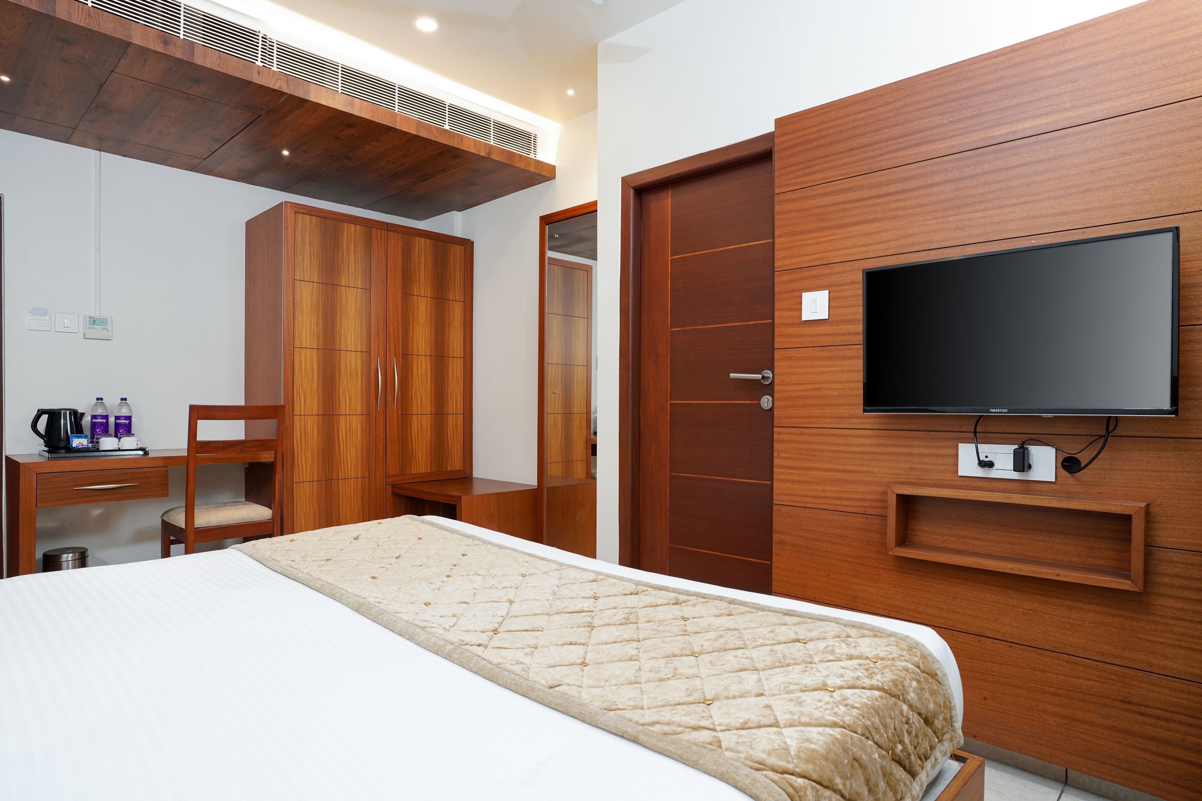 Una habitación de hotel con cama, armario de madera, escritorio con silla, televisor montado en la pared y una pequeña estantería.