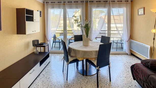 A dining area with a round table, chairs, a vase with flowers, and a view through large windows.