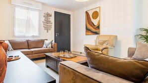 A modern living room with a brown leather sofa, a wooden coffee table, and a beige armchair. There is a black door, a window with sheer curtains, and a wall-mounted abstract painting. - Charming House with Garden, Close to Beaches & Guérande, Free WiFi (Assérac)