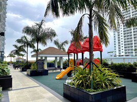 A children's playground with slides and a playhouse, surrounded by palm trees and modern buildings.