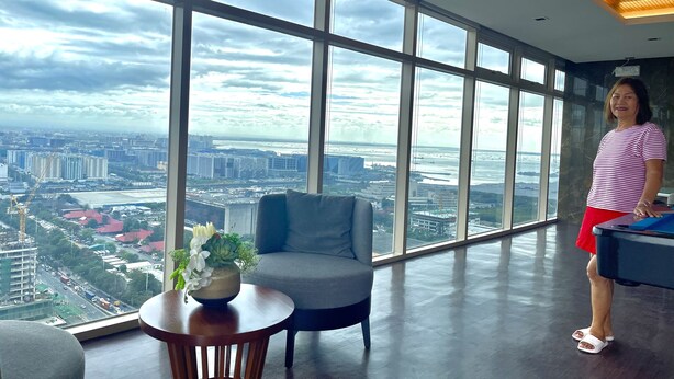 Une pièce spacieuse avec de grandes fenêtres donnant sur la ville, un fauteuil gris, une petite table en bois avec une plante et un bureau avec un ordinateur.