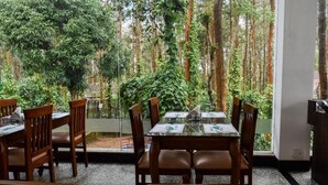 A dining area with a table and chairs, a view of a forest through a glass wall, and a door to the right. - Nexstay Coffee Grove Resort (Chikkamagaluru)