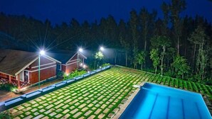 A swimming pool with a paved deck, surrounded by a well-maintained lawn and a row of trees. - Nexstay Coffee Grove Resort (Chikkamagaluru)