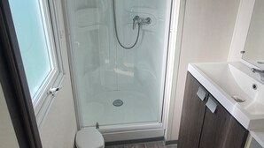 A modern bathroom with a glass shower enclosure, a white toilet, and a vanity with a mirror. - "mobil-home Sweet Home" : 32 m² 3 Chambres, 6 Personnes, au Calme Avec Jardin (Houlgate)