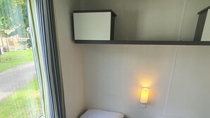 A compact room with two beds, a window with curtains, a bedside table, and a wall-mounted light. - "mobil-home Sweet Home" : 32 m² 3 Chambres, 6 Personnes, au Calme Avec Jardin (Houlgate)