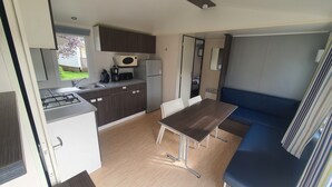 A compact kitchenette with a gas stove, microwave, and refrigerator. There is a dining table with chairs and a blue sofa. - "mobil-home Sweet Home" : 32 m² 3 Chambres, 6 Personnes, au Calme Avec Jardin (Houlgate)