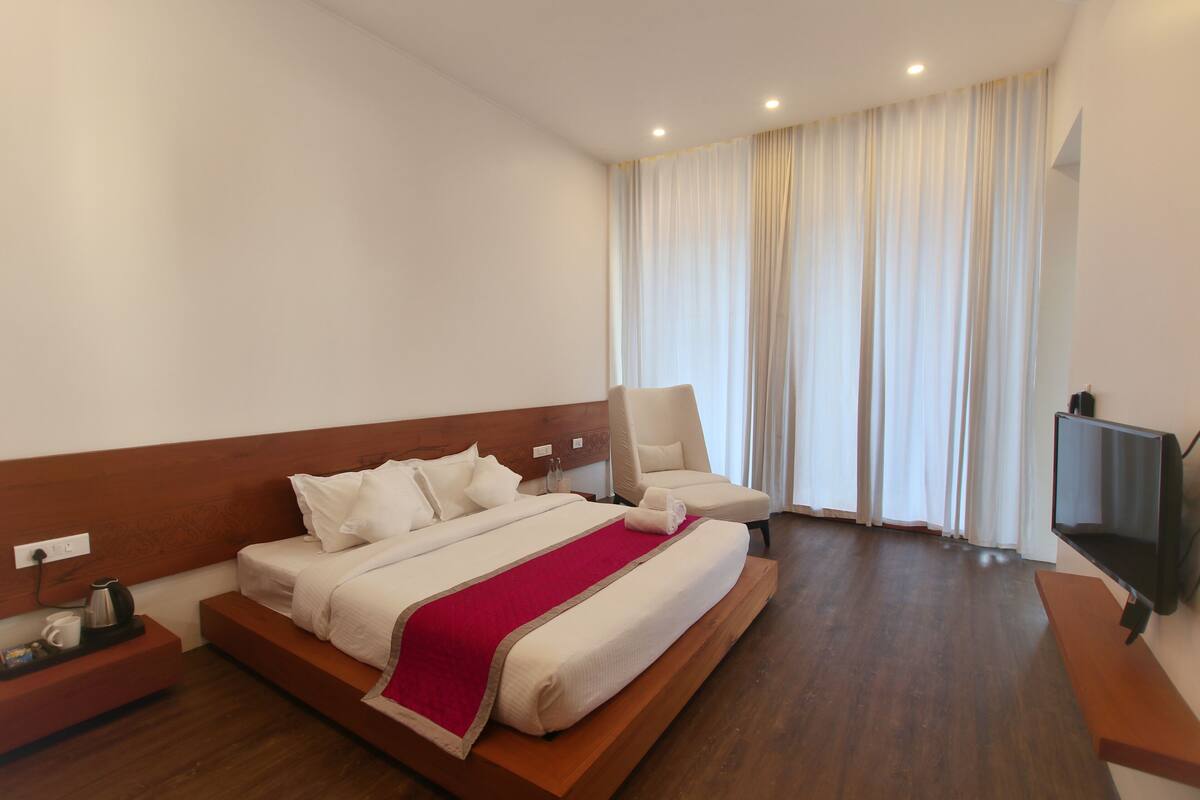 Ein Hotelzimmer mit einem großen Bett, einem Sessel, einem Fernseher und einem Fenster mit Vorhängen.