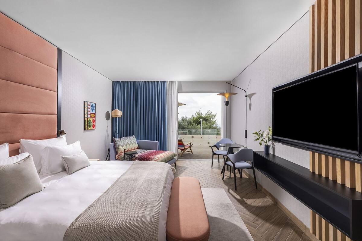 A modern hotel room with a large bed, a flat-screen TV, a seating area with a view, and a balcony.