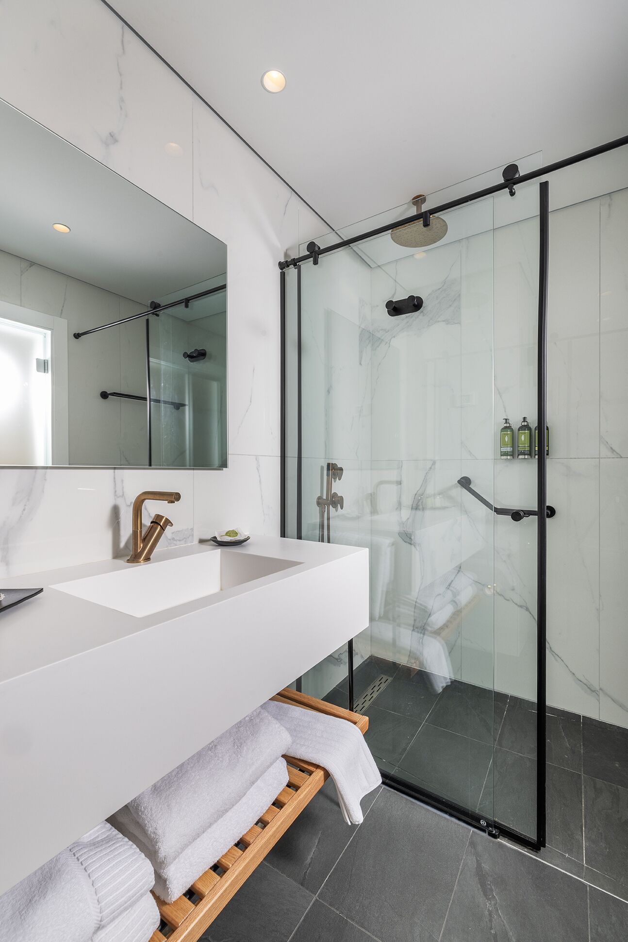 Un baño moderno con cabina de ducha de vidrio, tocador blanco con grifo dorado y banco de madera con toallas.