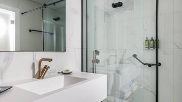 Un baño moderno con cabina de ducha de vidrio, tocador blanco con grifo dorado y banco de madera con toallas.