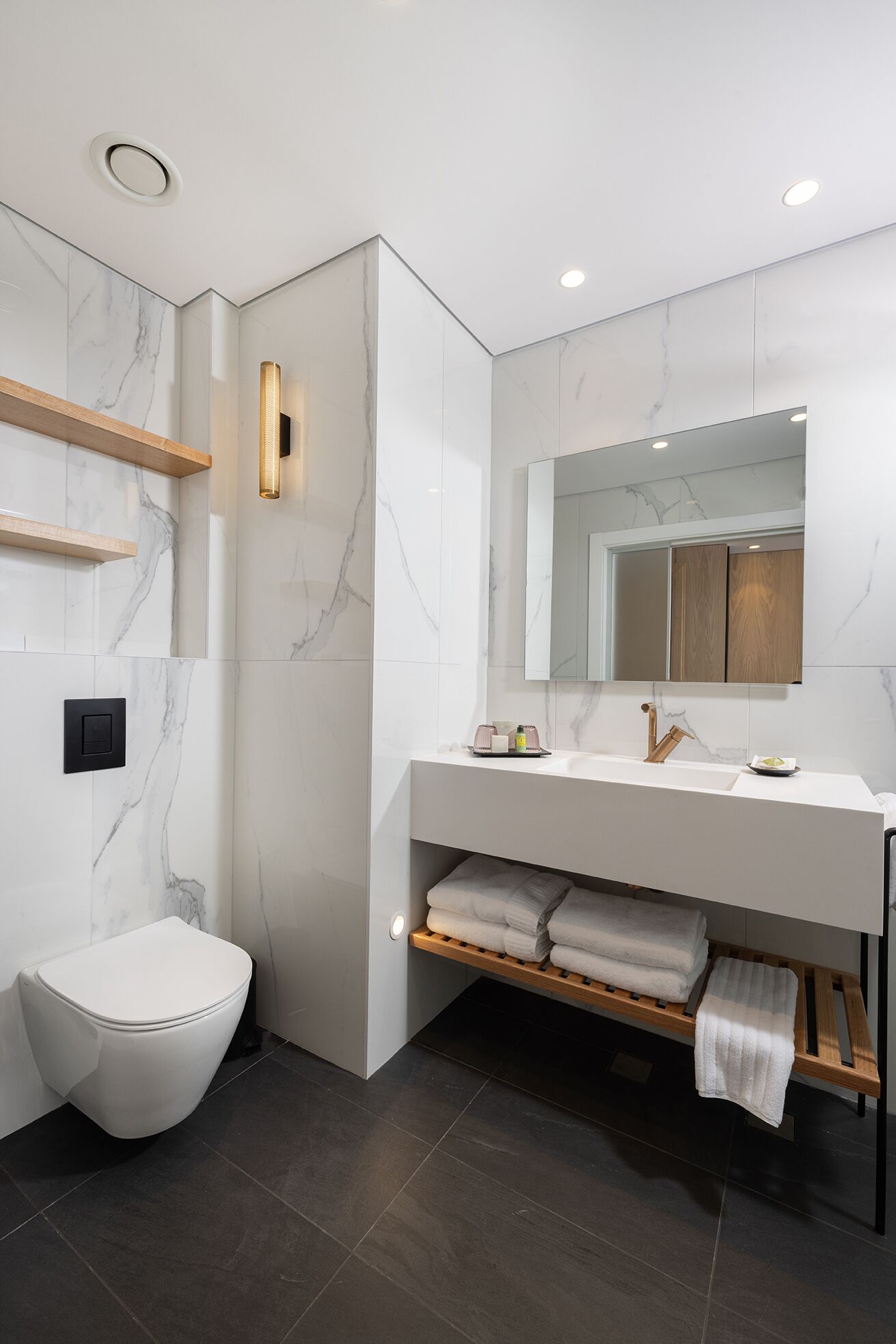 Une salle de bain moderne avec un mur en marbre blanc, un meuble-lavabo en bois, des toilettes et une étagère intégrée où sont rangées des serviettes.