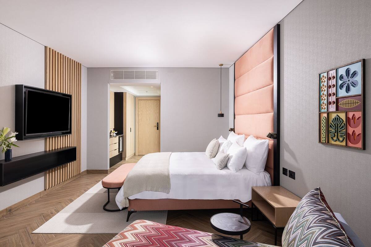 A modern hotel room with a large bed, a flat-screen TV mounted on the wall, a wooden floor, and a small seating area with a table and chairs.
