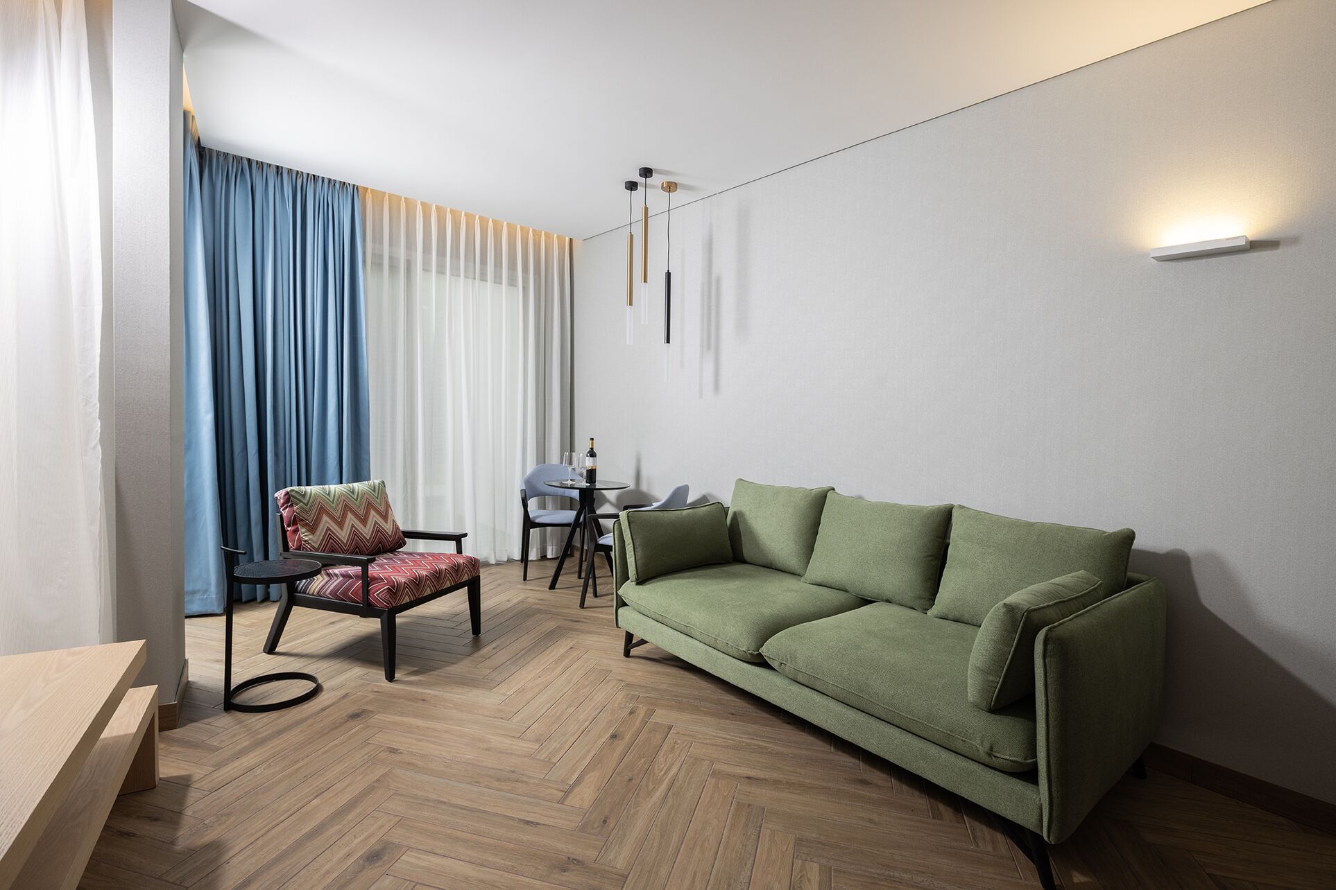 Una sala de estar moderna con un sofá verde, suelo de madera en patrón de espiga, una mesa redonda pequeña con sillas y una ventana con cortinas azules y blancas.